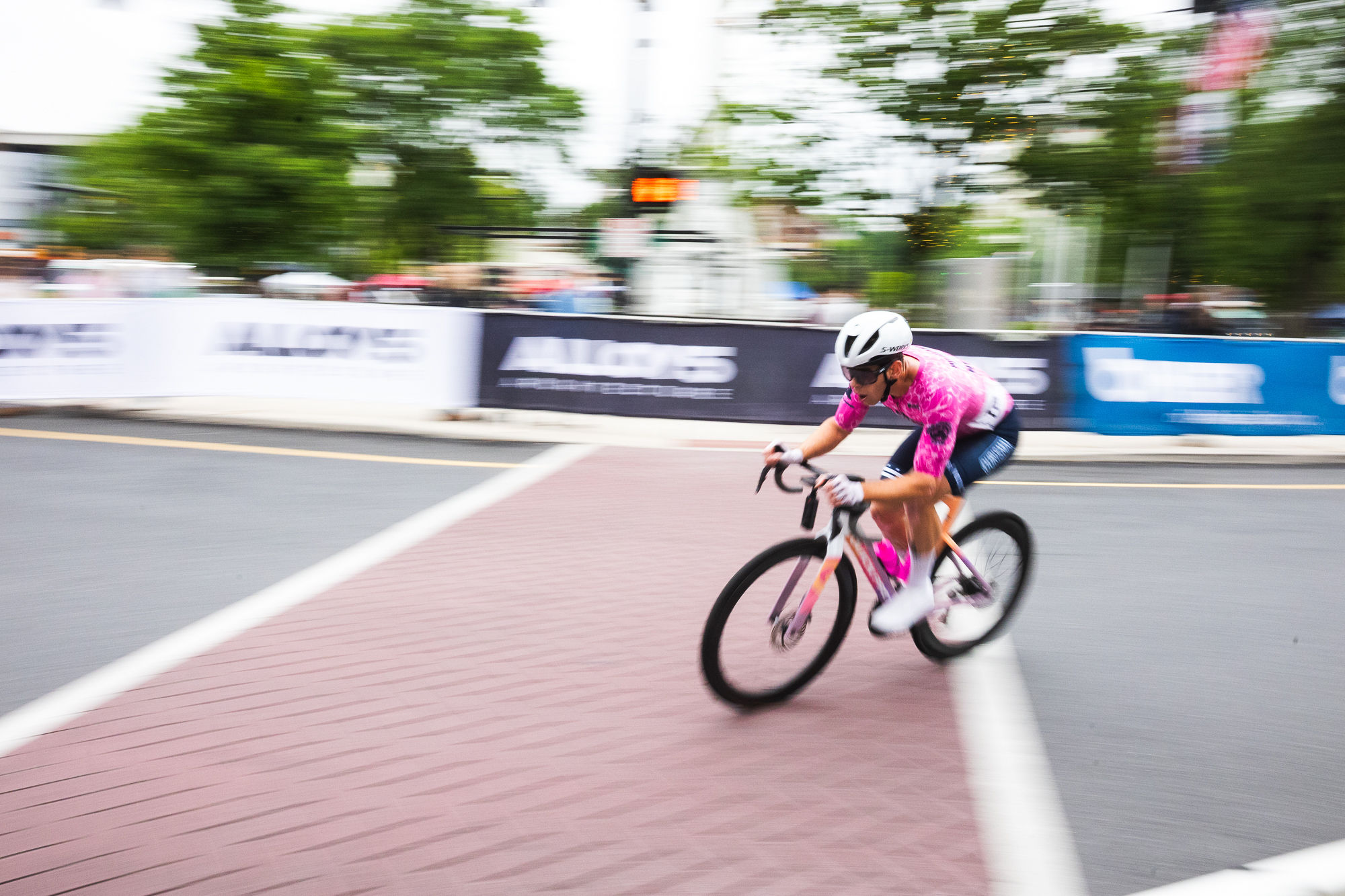 Rider in a custom pink Sportkraft jersey cornering at speed on the brick crosswalk of a criterium